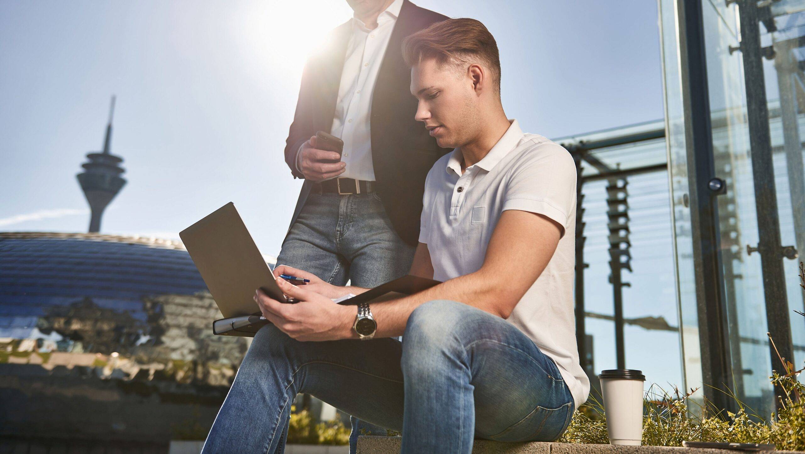 Ein junger und ein älterer Geschäftsmann arbeiten in der Stadt in der Sonne am Notebook und haben ein Meeting.